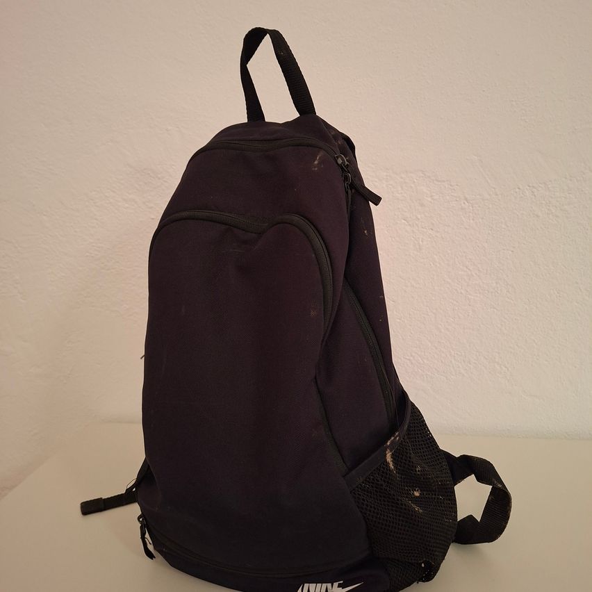 Ein schwarzer Rucksack mit Reißverschluss steht auf einer weißen Oberfläche. Der Rucksack hat zwei Reißverschlüsse und eine Netztasche vorne.