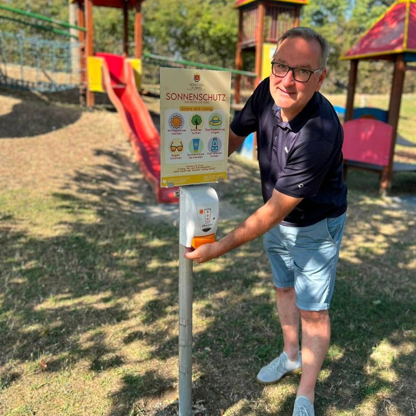 Ein Mann steht neben einem Sonnenschutzschild im Spielplatz und hält eine Flasche Sonnencreme in der Hand.