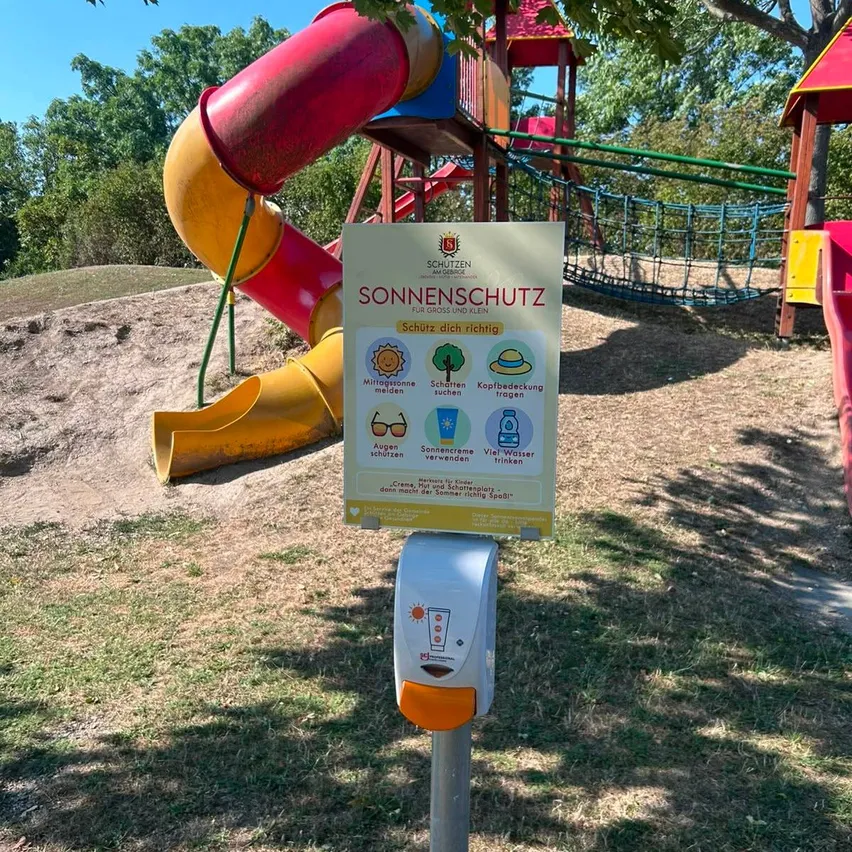 Ein Schild mit Sonnenschutztipps steht auf einem Pfosten in einem Spielplatz. Dahinter befindet sich eine Rutsche und eine Kletterstruktur. Das Schild hat einen gelben Hintergrund mit weißen Texten und Symbolen.