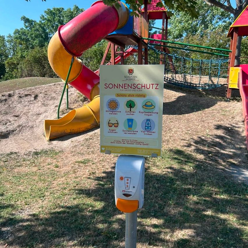 Ein Schild mit Sonnenschutztipps steht auf einem Pfosten in einem Spielplatz. Dahinter befindet sich eine Rutsche und eine Kletterstruktur. Das Schild hat einen gelben Hintergrund mit weißen Texten und Symbolen.