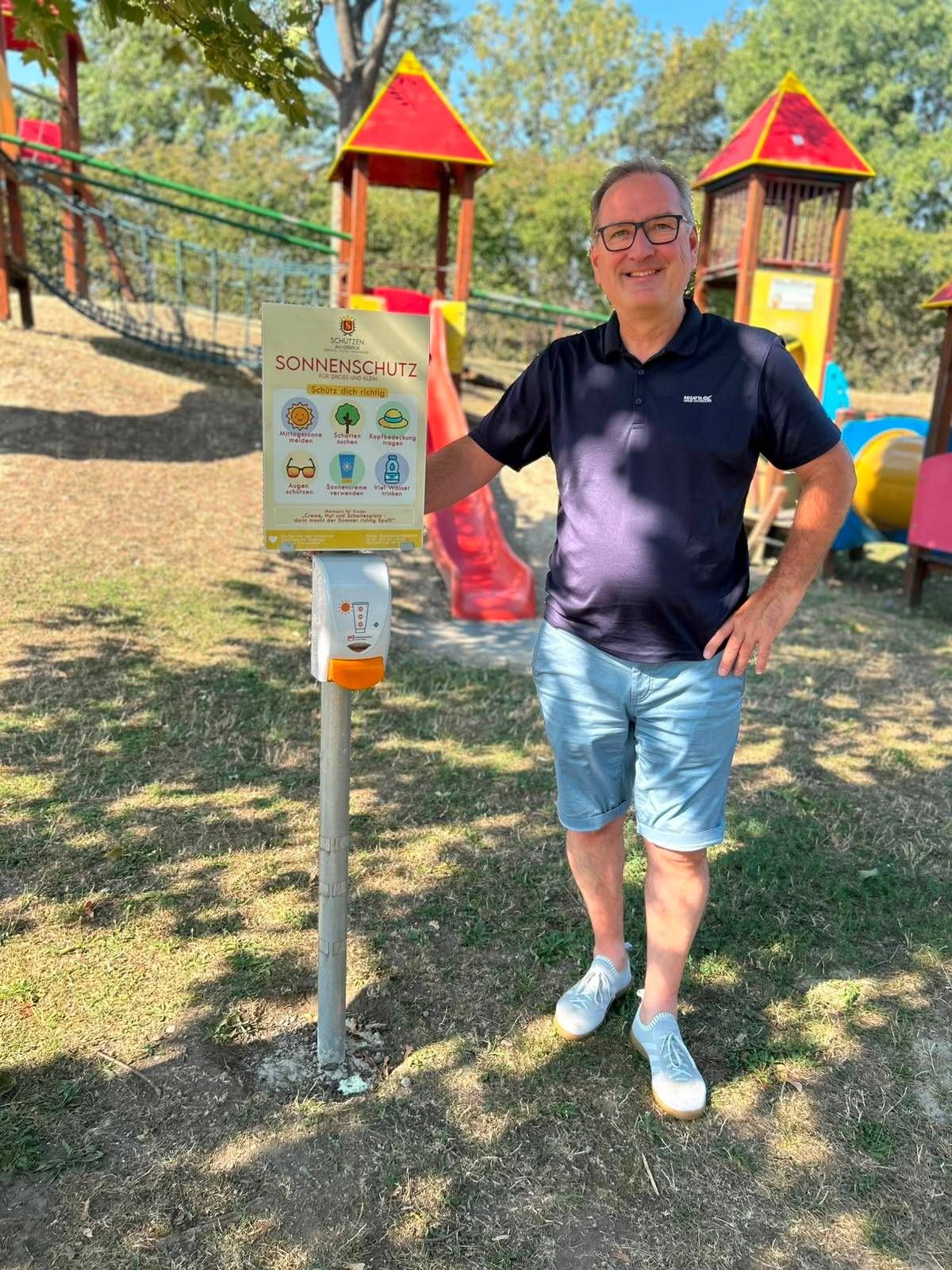 Ein Mann steht neben einem Schild mit Sonnenschutztipps und einem Handdesinfektionsspender auf einem Spielplatz.