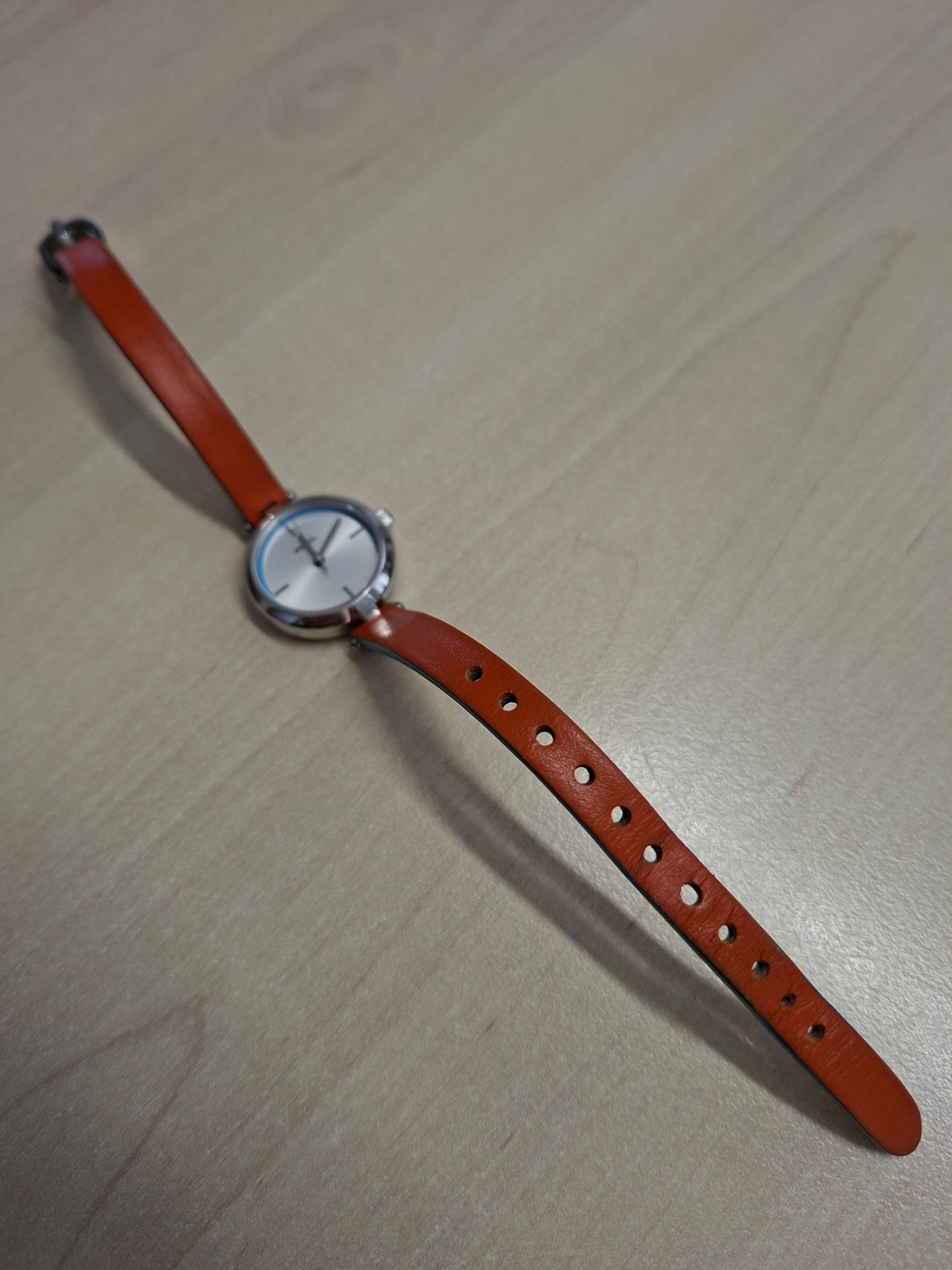 Eine kleine Armbanduhr mit einem braunen Lederband und einem silberfarbenen Zifferblatt liegt auf einer Holzoberfläche.