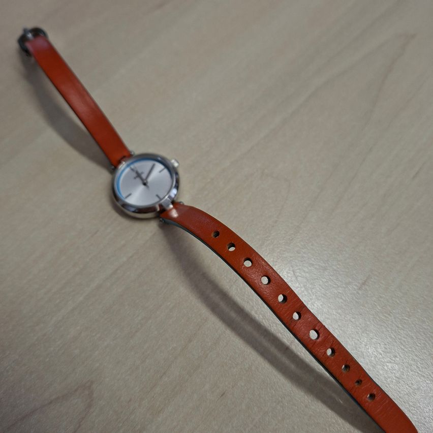 Eine kleine Armbanduhr mit einem braunen Lederband und einem silberfarbenen Zifferblatt liegt auf einer Holzoberfläche.