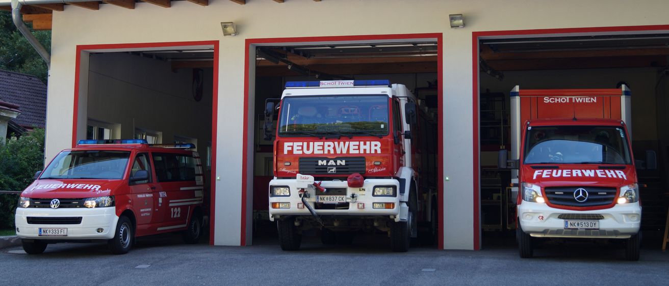 Ein Feuerwehrauto steht in einer Garage zusammen mit zwei anderen Einsatzfahrzeugen. Das Feuerwehrauto ist rot mit dem Wort 'Feuerwehr' auf der Seite. Das Gebäude hat rote und weiße Akzente.