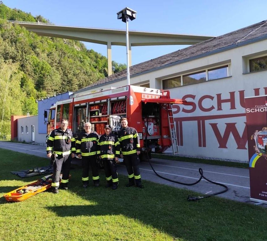 Vier Feuerwehrleute stehen vor einem Feuerwehrauto mit offener Tür in der Nähe eines Gebäudes mit einem Schild, das SCHW besagt.
