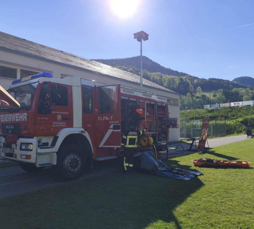 Ein roter Feuerwehrwagen mit einem Feuerwehrmann draußen, eine Trage und ein Feuerlöscher in der Nähe. Der Wagen ist in der Nähe eines Gebäudes mit einem geneigten Dach geparkt. Berge sind in der Ferne.