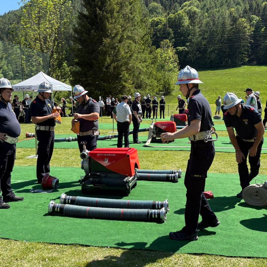 Feuerwehrleute in Uniform bereiten Ausrüstung für eine Trainingsübung auf einem grünen Feld vor, mit Zuschauern im Hintergrund.