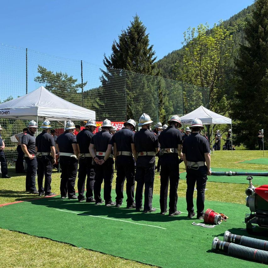 Eine Gruppe von Feuerwehrleuten in Uniform steht in einer Reihe auf einem grünen Feld. Sie tragen Helme und Gürtel. Zelte im Hintergrund mit einem Zaun.