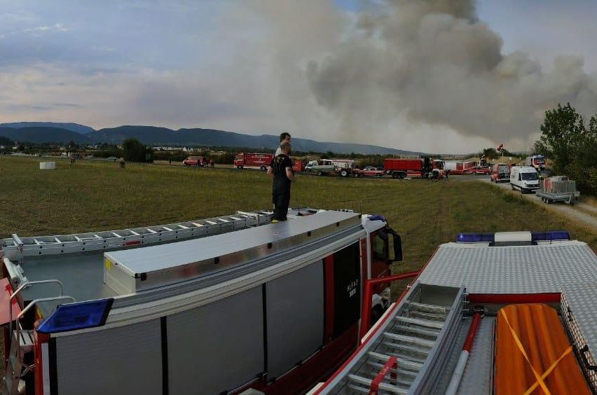 Feuerwehrleute sind am Brandort, mit aufsteigendem Rauch im Hintergrund. Mehrere Feuerwehrwagen sind auf einem Feld geparkt, mit Bergen in der Ferne.