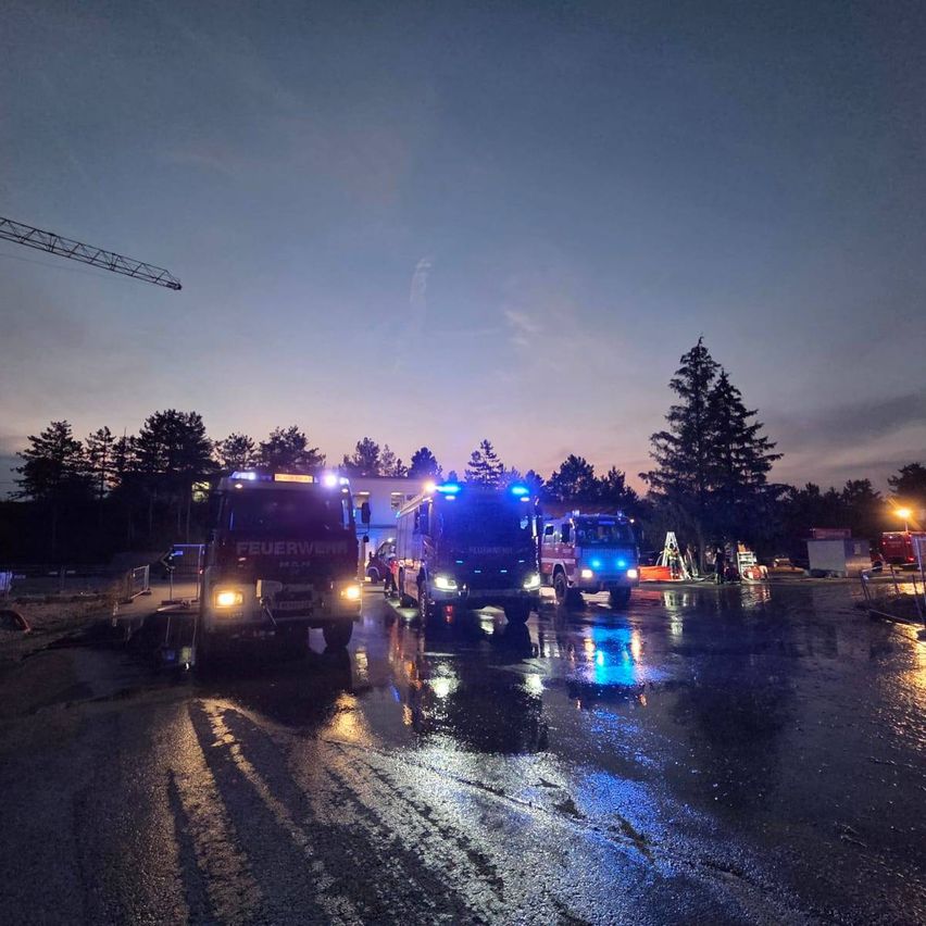 Drei Feuerwehrfahrzeuge stehen bei Nacht auf einer nassen Straße mit eingeschalteten Lichtern. Im Hintergrund sind Bäume und ein Kran zu sehen.