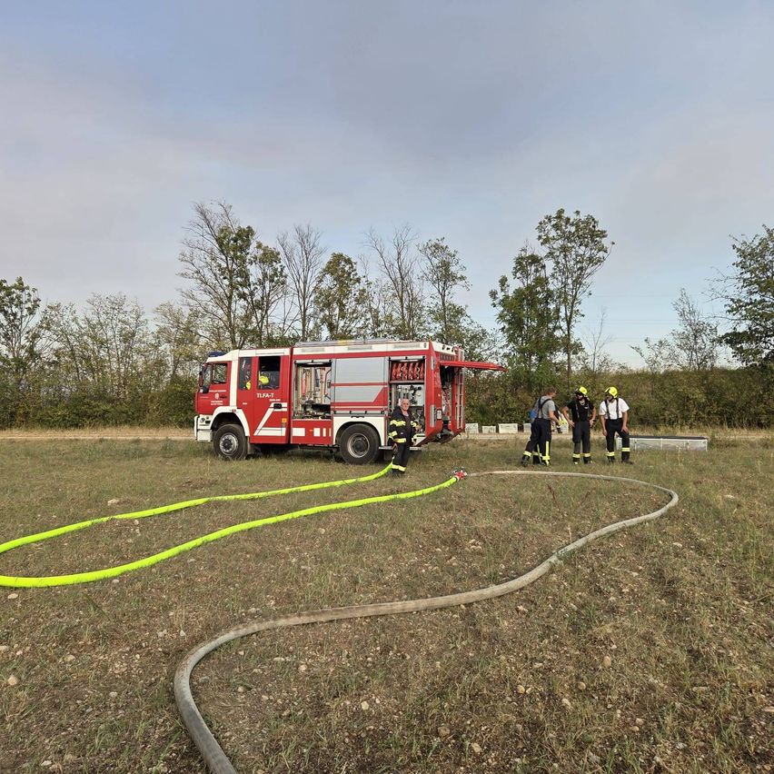 Ein roter Feuerwehrwagen steht auf einem Grasfeld, um ihn herum stehen Feuerwehrleute. Ein gelber Schlauch liegt auf dem Boden.
