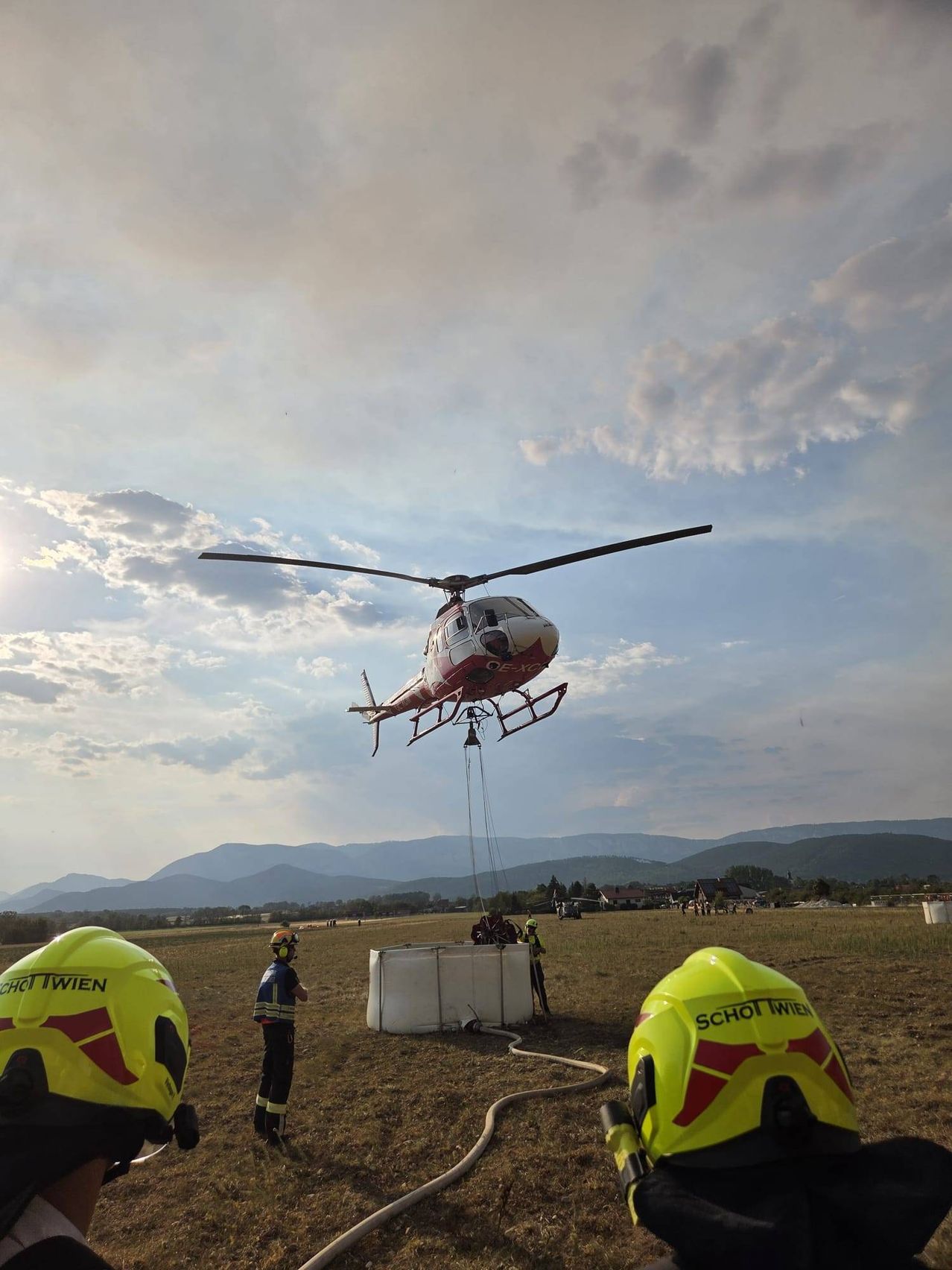 Ein roter und weißer Hubschrauber befindet sich in der Luft und wird durch ein Seil abgesenkt. Mehrere Personen in gelben Helmen stehen am Boden, eine hält einen Schlauch. Die Szene spielt sich auf einem Feld mit Bergen im Hintergrund ab.
