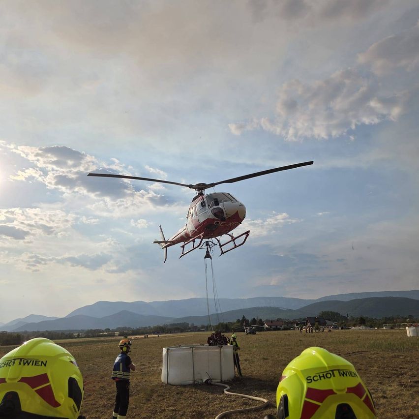 Ein roter und weißer Hubschrauber befindet sich in der Luft und wird durch ein Seil abgesenkt. Mehrere Personen in gelben Helmen stehen am Boden, eine hält einen Schlauch. Die Szene spielt sich auf einem Feld mit Bergen im Hintergrund ab.