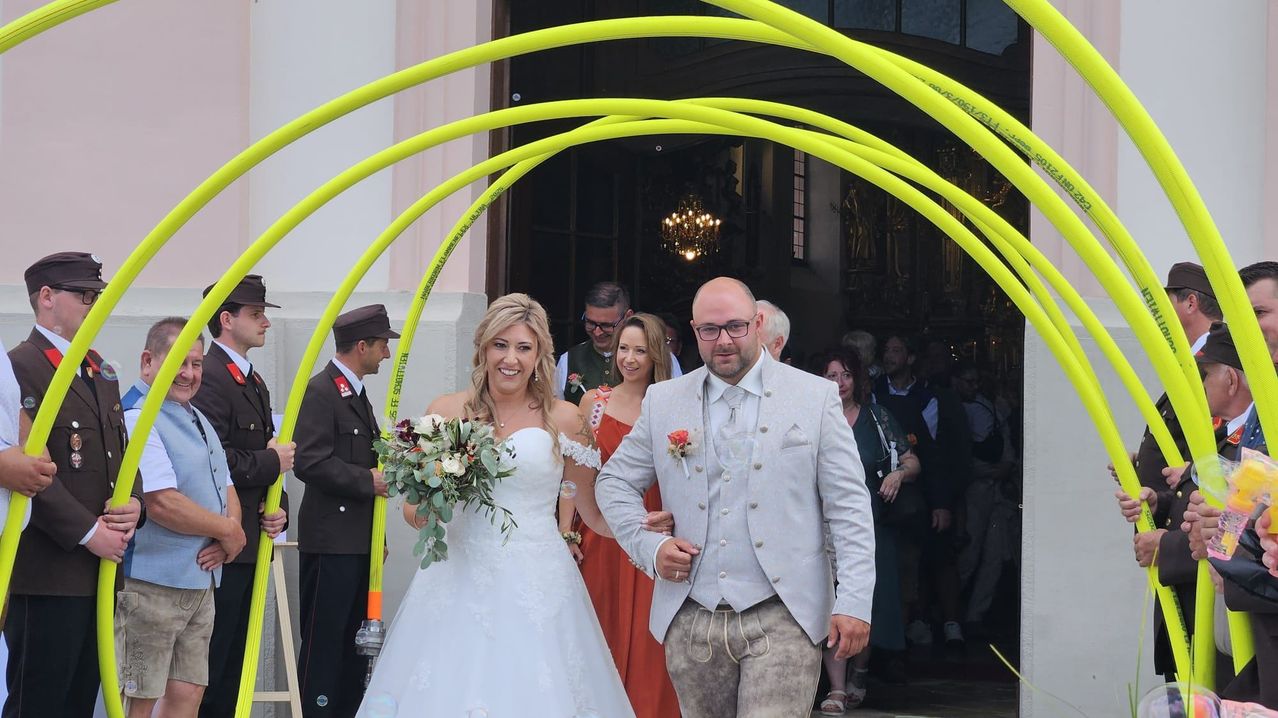 Eine Braut in einem weißen Hochzeitskleid und ein Bräutigam in einem grauen Anzug gehen durch einen gelben Bogen bei einer Hochzeitszeremonie.