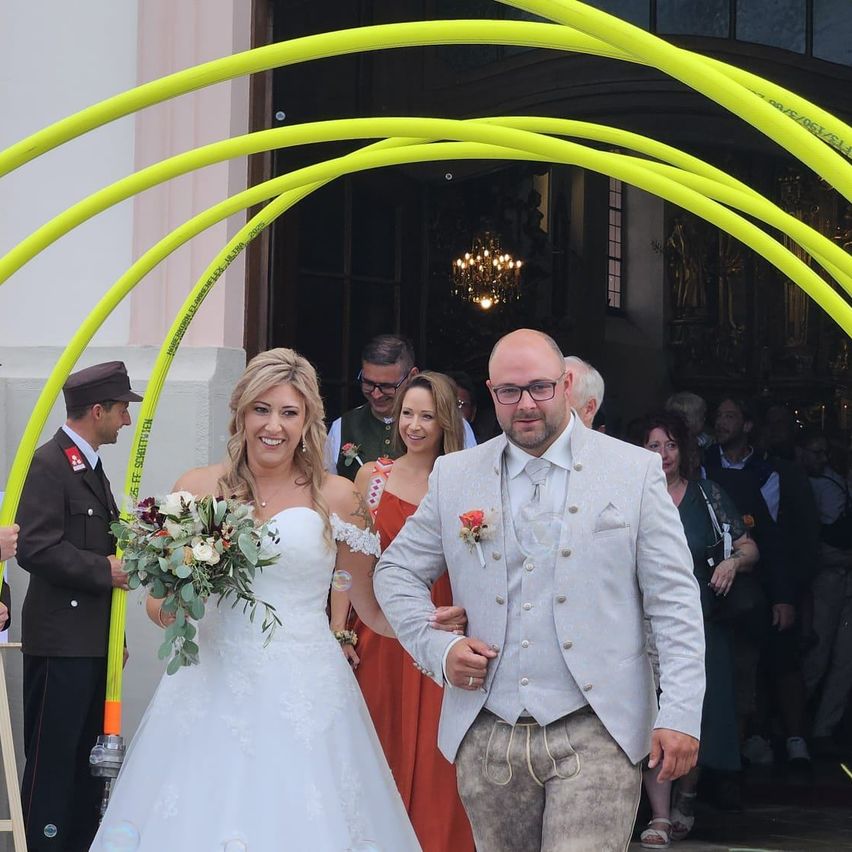 Eine Braut in einem weißen Hochzeitskleid und ein Bräutigam in einem grauen Anzug gehen durch einen gelben Bogen bei einer Hochzeitszeremonie.