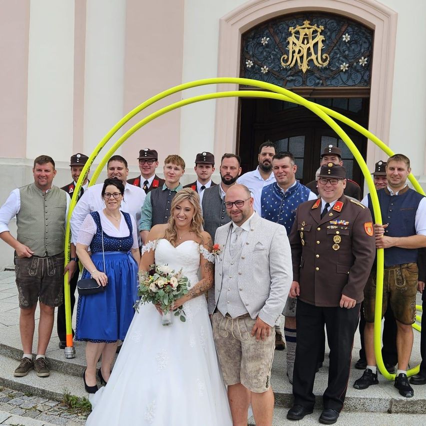 Eine Gruppe von Menschen in traditioneller deutscher Kleidung, einschließlich einer Braut in einem weißen Hochzeitskleid, posiert für ein Foto vor einem Gebäude.