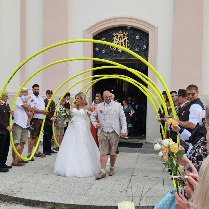 Ein Hochzeitspaar geht durch einen Bogen aus gelben Röhren, umgeben von Gästen mit Blumen. Die Braut trägt ein weißes Kleid, der Bräutigam ein helles Kostüm. Sie sind vor einer Kirche.