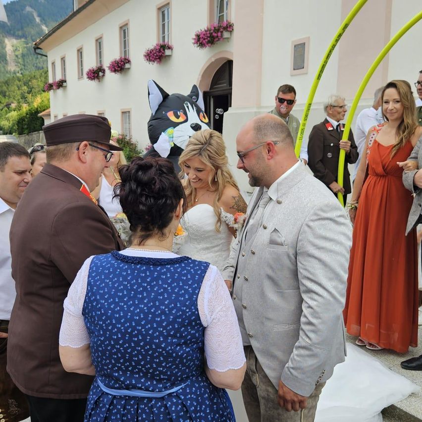 Eine Hochzeitszeremonie im Freien mit einem großen Katzenballon im Hintergrund. Ein Brautpaar steht vor Gästen, eine Frau in einem blauen Kleid und ein Mann in einem braunen Anzug schauen sie an.