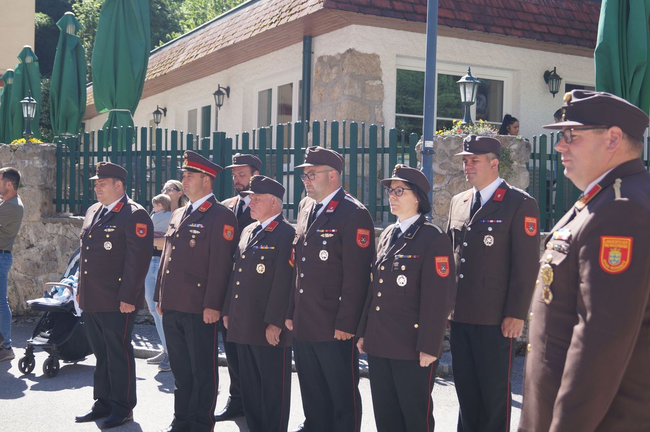 Eine Gruppe uniformierter Personen, möglicherweise eine Militäreinheit, steht vor einem Gebäude. Sie tragen Mützen, Medaillen und Brillen. Das Gebäude hat grüne Zäune, Fenster und eine Lampe.