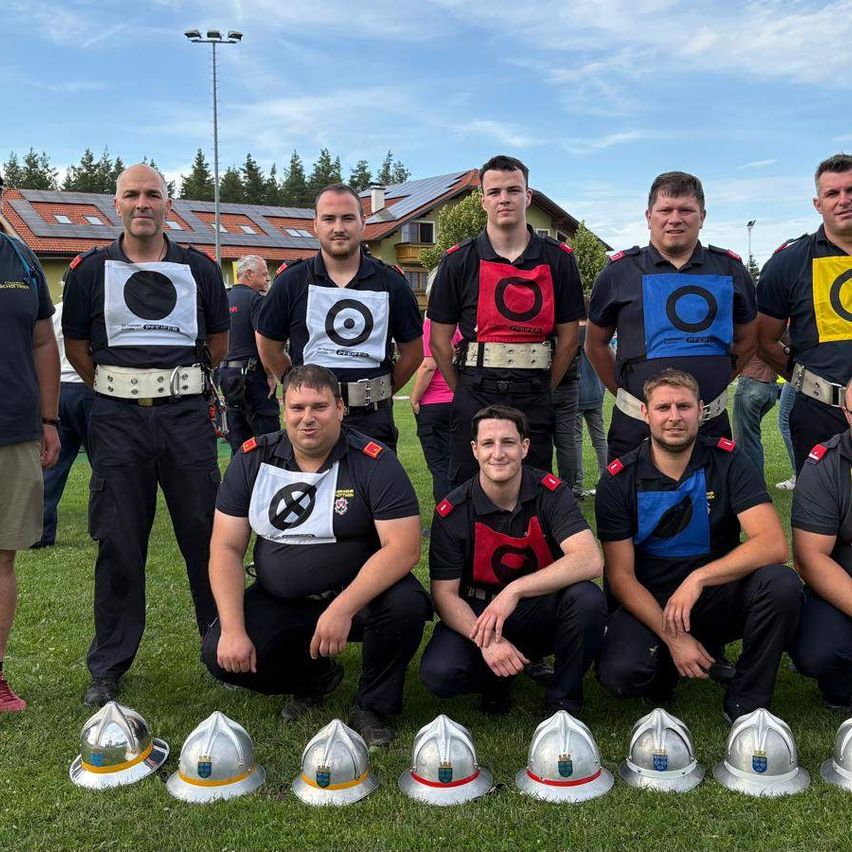 Eine Gruppe von Feuerwehrleuten in Uniform posiert für ein Foto auf einem Feld, mit Helmen, die davor aufgereiht sind.