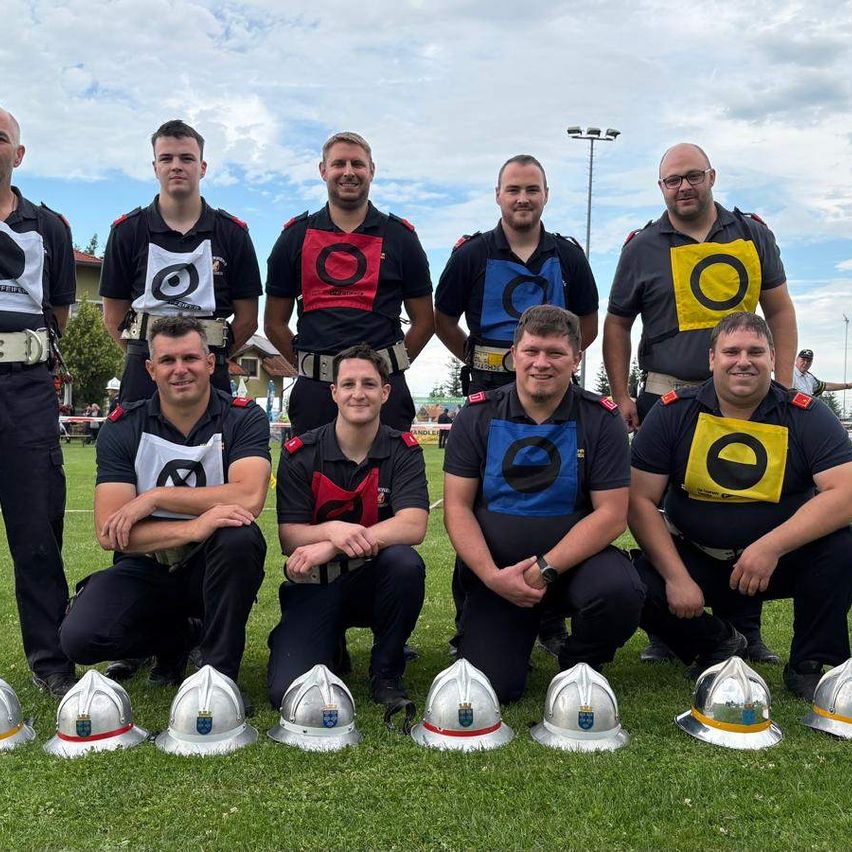 Eine Gruppe Feuerwehrleute posiert für ein Foto, trägt Uniformen und Helme mit farbigen Aufnähern. Sie befinden sich auf einem Rasenfeld mit Helmen davor.