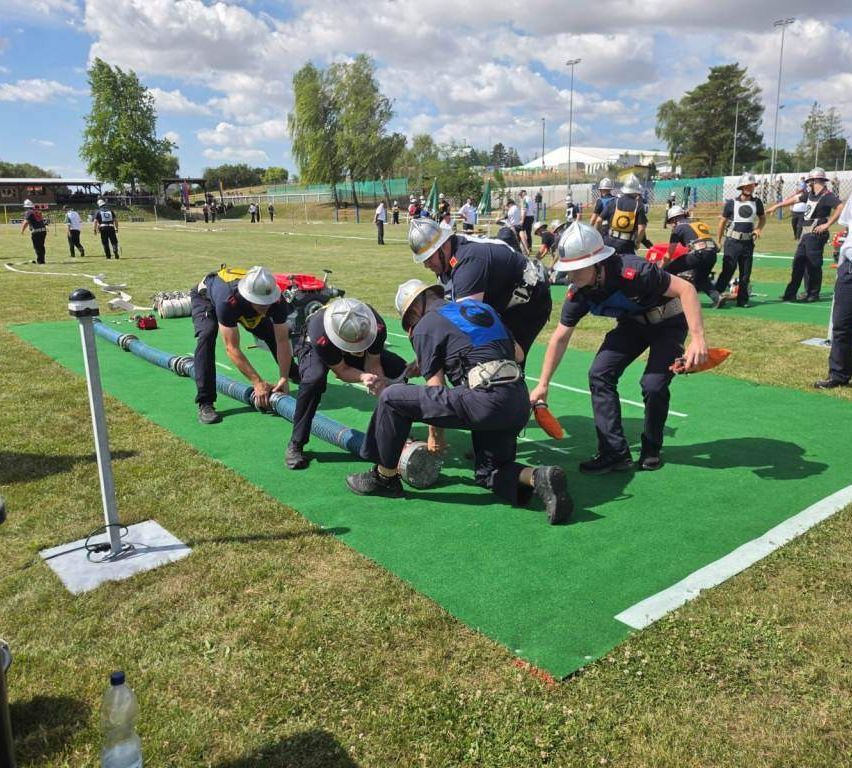 Eine Gruppe Feuerwehrleute in Helmen übt auf einem grünen Feld mit einem Schlauch und anderen Geräten. Zuschauer beobachten von hinten.