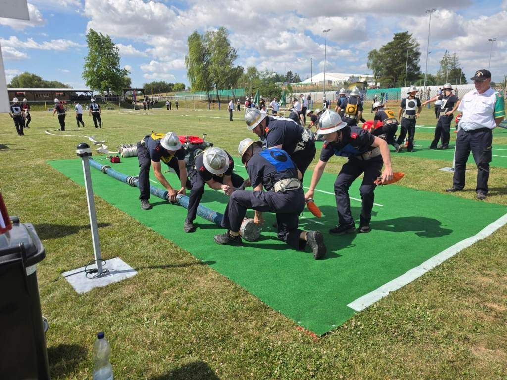 A group of firefighters in helmets are practicing on a green field, handling a hose and other equipment. Spectators are watching from behind.
