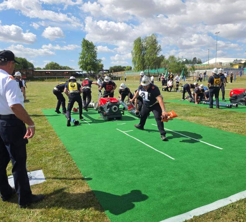 Several firefighters are participating in a training exercise on a green field with white lines. Some are wearing helmets, and one holds a bag. A person in the background is observing.