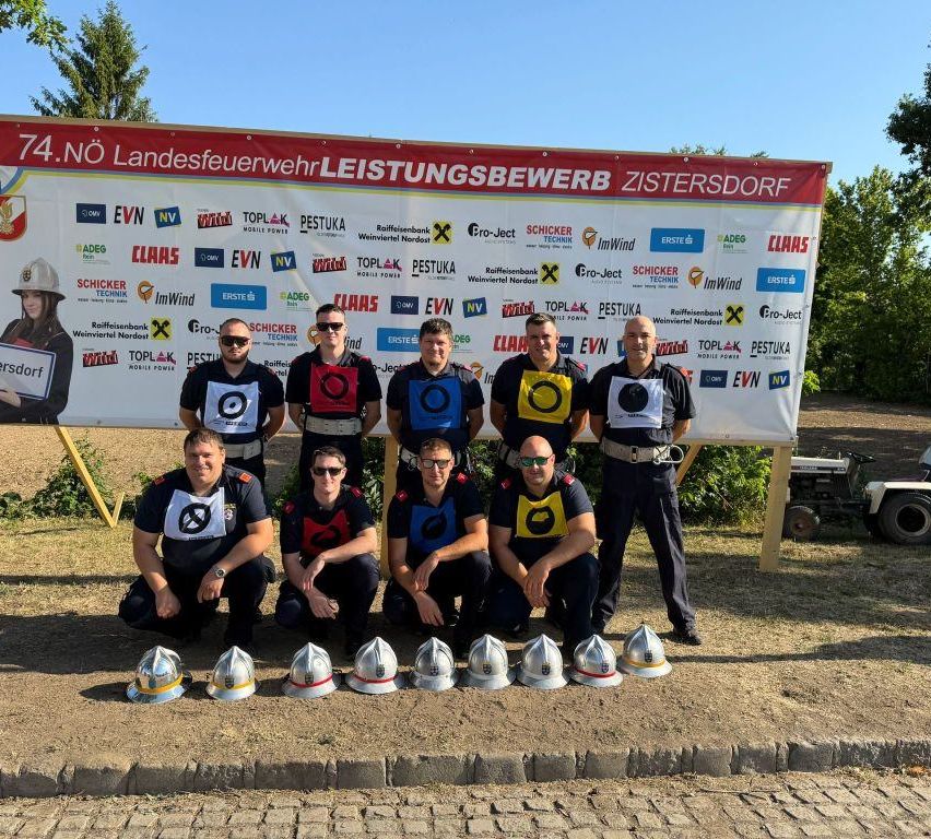 A group of firefighters in uniform pose for a photo with helmets in front of a banner that reads 'No. 4 Landesfeuerwehr Leistungsbewerb Zistersdorf.'