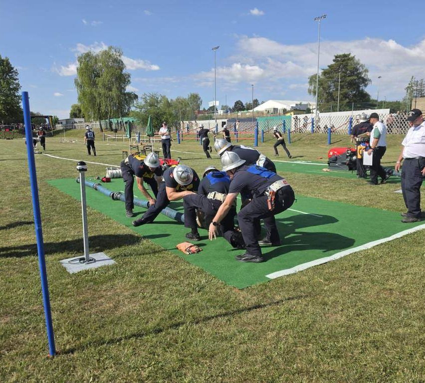 Firefighters practice a hose-laying exercise on a green field, wearing helmets and uniforms, with a crowd watching from afar.