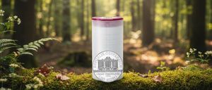 Ein weißer Plastikbecher mit rotem Deckel steht in einem Wald und zeigt eine 150-Euro-Münze mit einem Gebäudedesign.