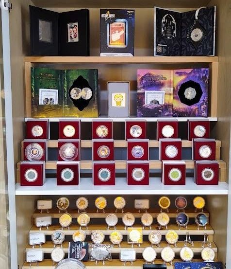 Ein Vitrinenschrank mit mehreren Regalen, gefüllt mit Münzen und Gegenständen in roten und schwarzen Schachteln. Das oberste Regal hat größere Schachteln mit 'Star Trek' und anderen Etiketten. Die mittleren Regale haben Reihen von Münzen in verschiedenen Farben. Das unterste Regal hat größere Münzen, einige mit Etiketten.