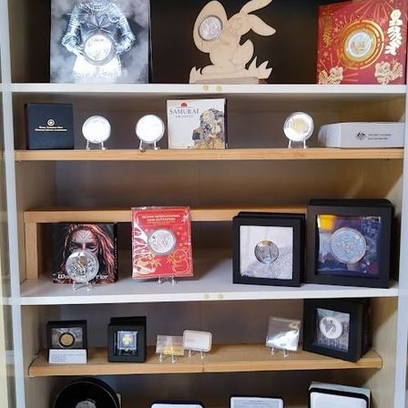 Ein holzfarbenes Regal zeigt verschiedene gerahmte Bilder, Münzen und Schachteln. Eine Münze zeigt einen Samurai, eine andere einen Hasen und andere verschiedene Designs.