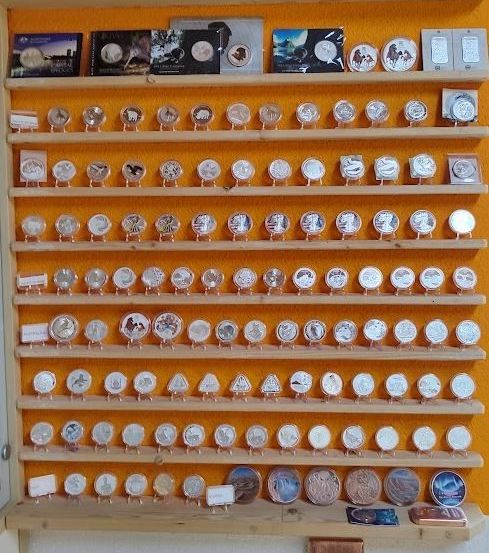 Ein holzfarbener Regal zeigt eine Sammlung von Münzen und Karten auf orangefarbenem Hintergrund. Die Münzen sind auf Ständern ausgestellt, während sich die Karten im obersten Regal befinden. Die Karten zeigen Bilder von Tieren und Menschen.