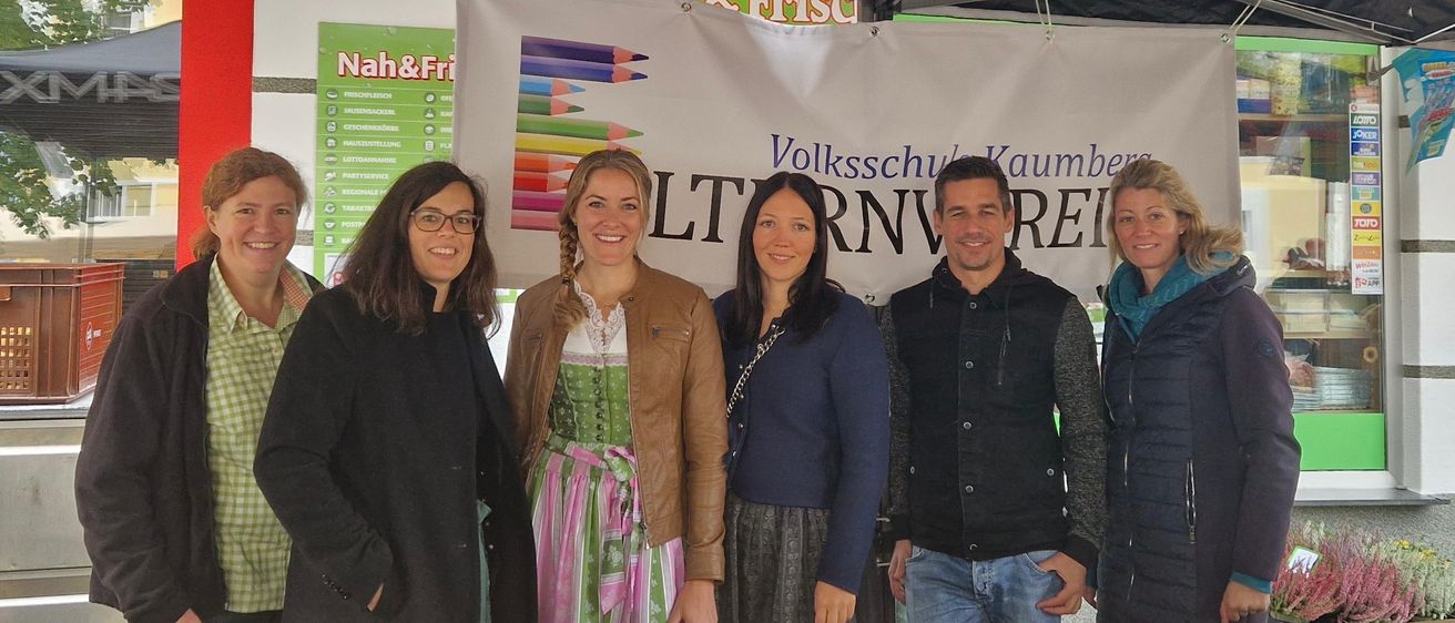 Fünf Personen stehen vor einem Banner. Zwei Frauen tragen traditionelle Kleidung. Auf dem Banner stehen 'Volksschule Kaumberg' und 'LTRNWREI'. Die Leute lächeln.