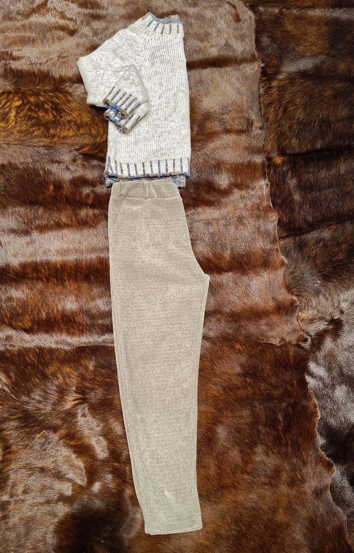 Ein Paar hellgrauer Hosen mit einem gefalteten weißen Pullover auf einer braunen Pelzoberfläche.