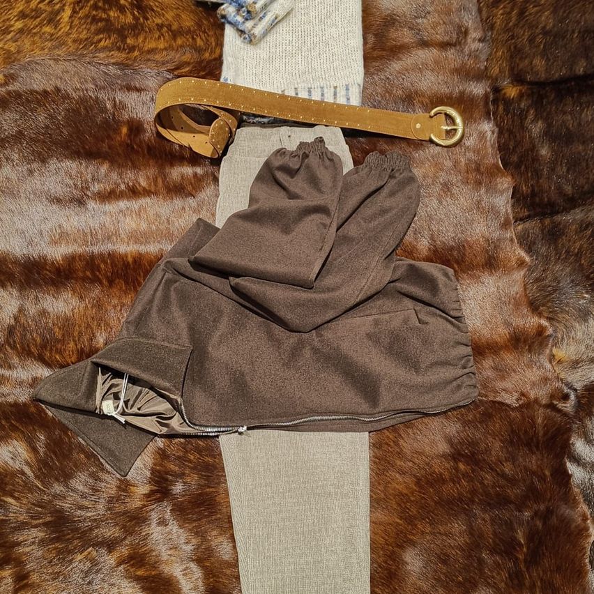 Ein brauner Gürtel, gefaltete Hosen und ein graues Kleidungsstück liegen auf einem braunen Pelzteppich.