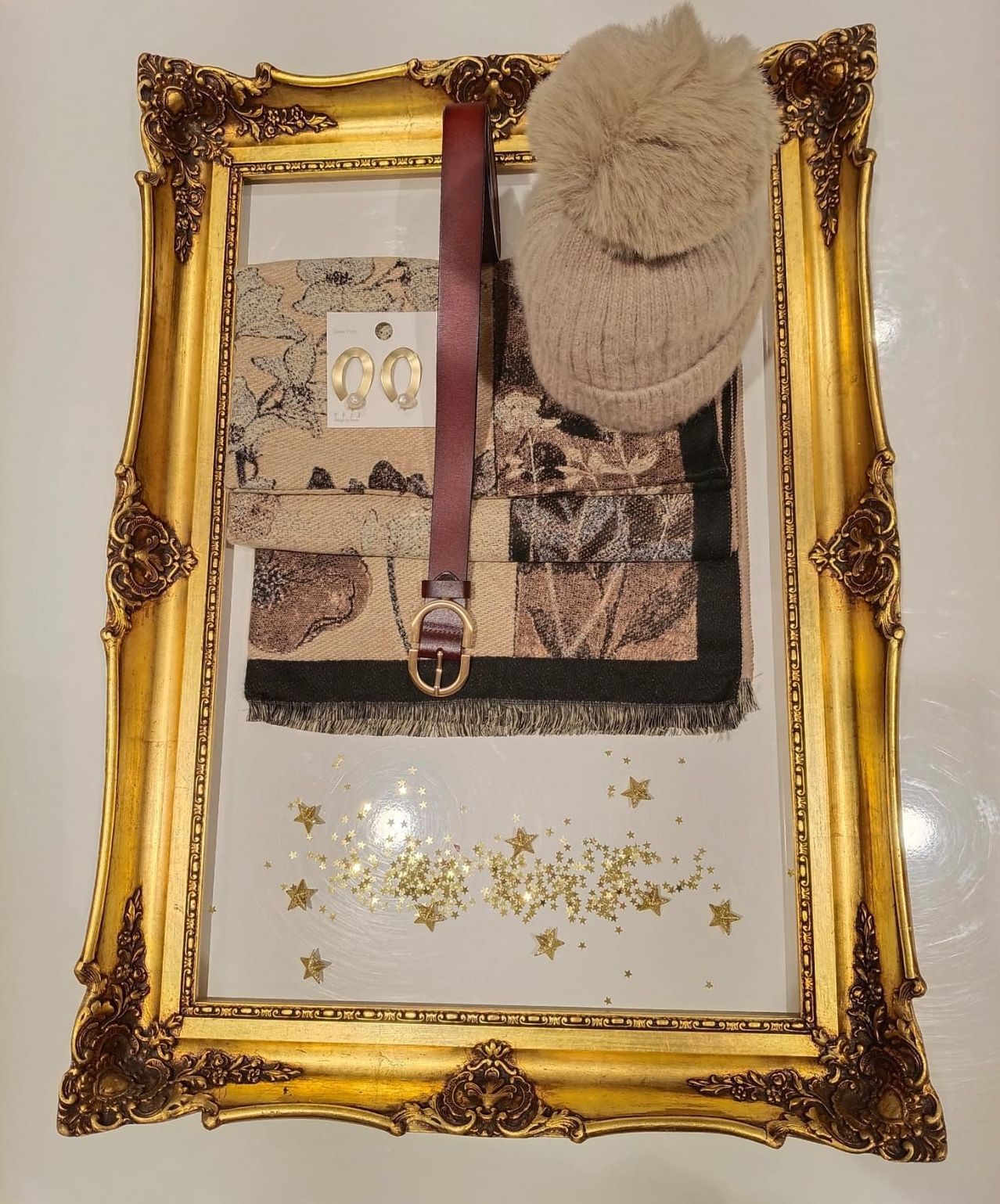 Ein goldener Rahmen zeigt ein Winterensemble mit einer Fellmütze, einem Schal und einem Gürtel. Die Gegenstände sind auf einem weißen Hintergrund angeordnet, mit goldenen Sternen darum verstreut.