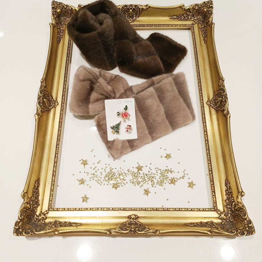 Ein goldener Rahmen mit weißem Hintergrund enthält drei braune Pelzartikel. Ein Pelzschal ist gefaltet und zwei Pelzumhänge sind darauf gelegt. Eine kleine Karte mit Weihnachtsdekorationen befindet sich in der Mitte.