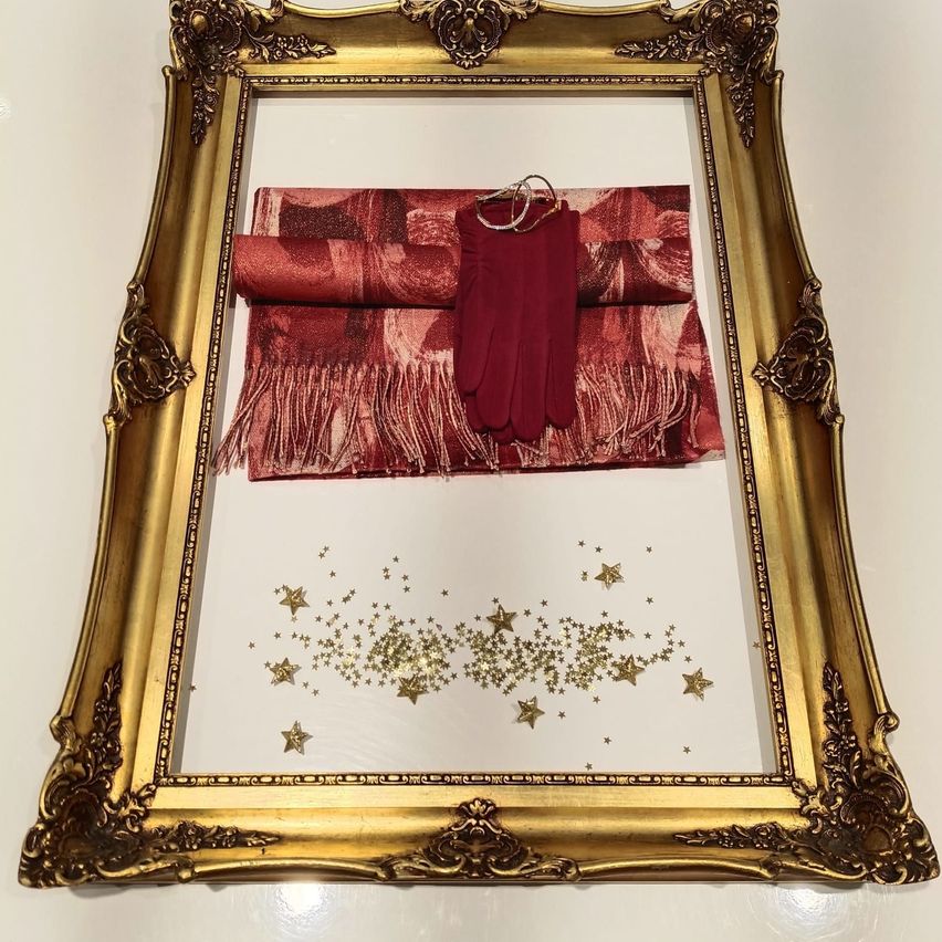 Ein goldfarbener Spiegelrahmen zeigt einen roten Schal, Handschuhe und einen Kleiderbügel. Der Spiegel ist mit goldenen Sternen verziert.