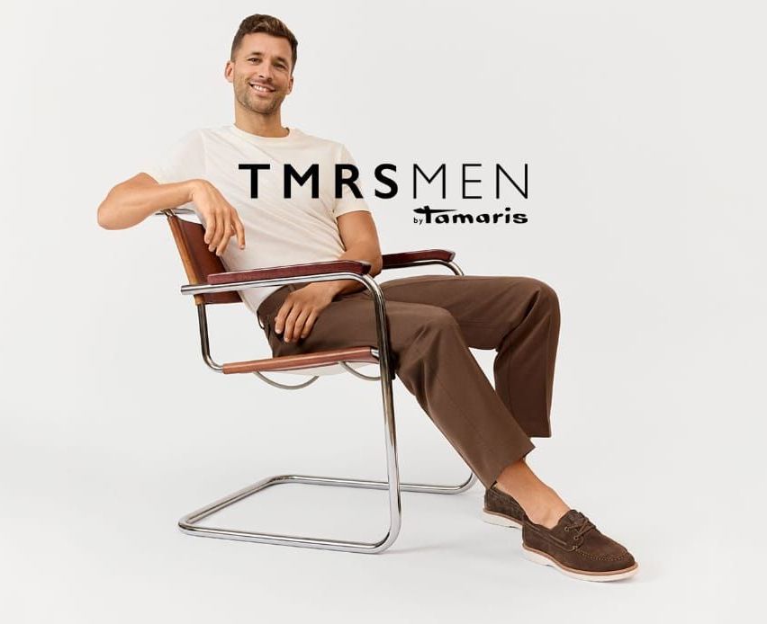 Ein lächelnder Mann, der ein weißes TMRSMENS by Tamaris T-Shirt und braune Hosen trägt, sitzt auf einem Stuhl. Er hält die Rückenlehne mit der linken Hand und die rechte Hand liegt auf dem Schoß. Er schaut nach links.