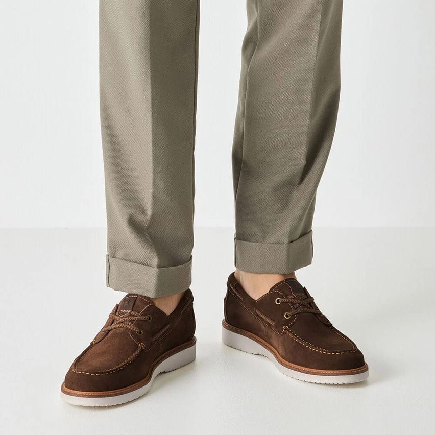 Eine Person in beigefarbenen Hosen und braunen Leder-Loafern mit weißen Sohlen steht auf einer weißen Oberfläche.