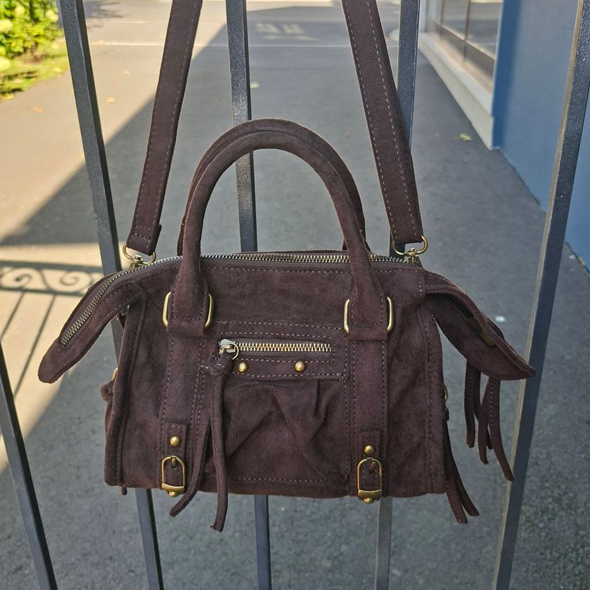Eine braune Wildlederhandtasche mit goldenen Beschlägen hängt an einem Metallzaun. Die Tasche hat einen Reißverschluss und einen gefransten Boden.