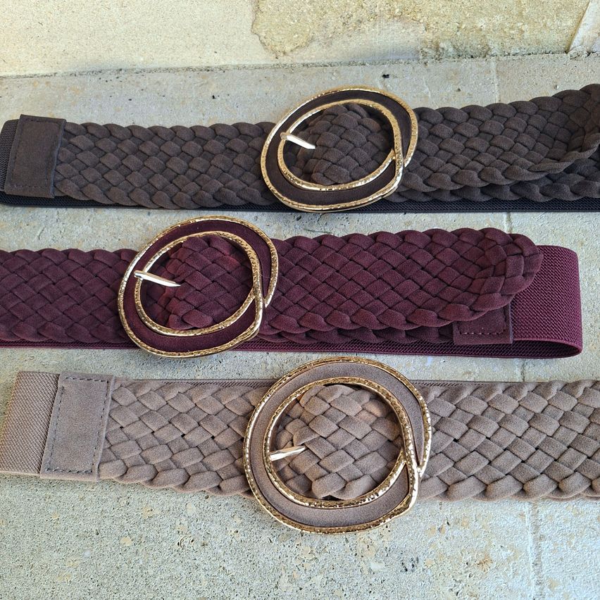 Drei Gürtel mit gewebten Texturen und goldenen Schnallen, auf einer Betonoberfläche ausgestellt. Die Gürtel sind in verschiedenen Farben: braun, bordeaux und beige.