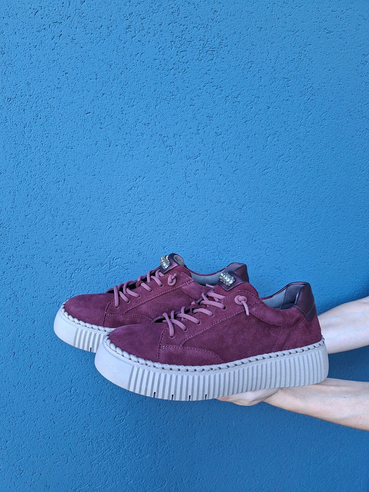 Ein Paar lila Wildleder-Sneaker mit weißen Sohlen und rosa Schnürsenkeln wird vor einem blauen Hintergrund gehalten.