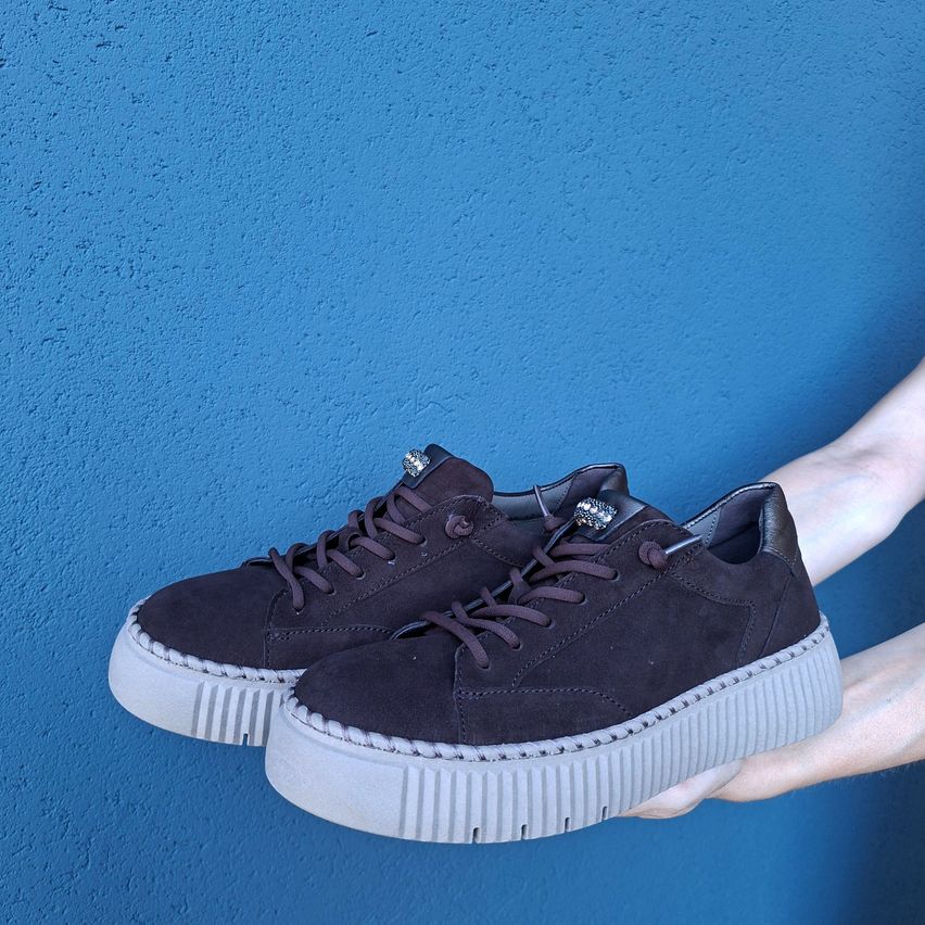 Ein Paar schwarze Wildleder-Plattform-Sneaker mit silbernen Akzenten, die von einer Hand vor einem blauen Hintergrund gehalten werden.
