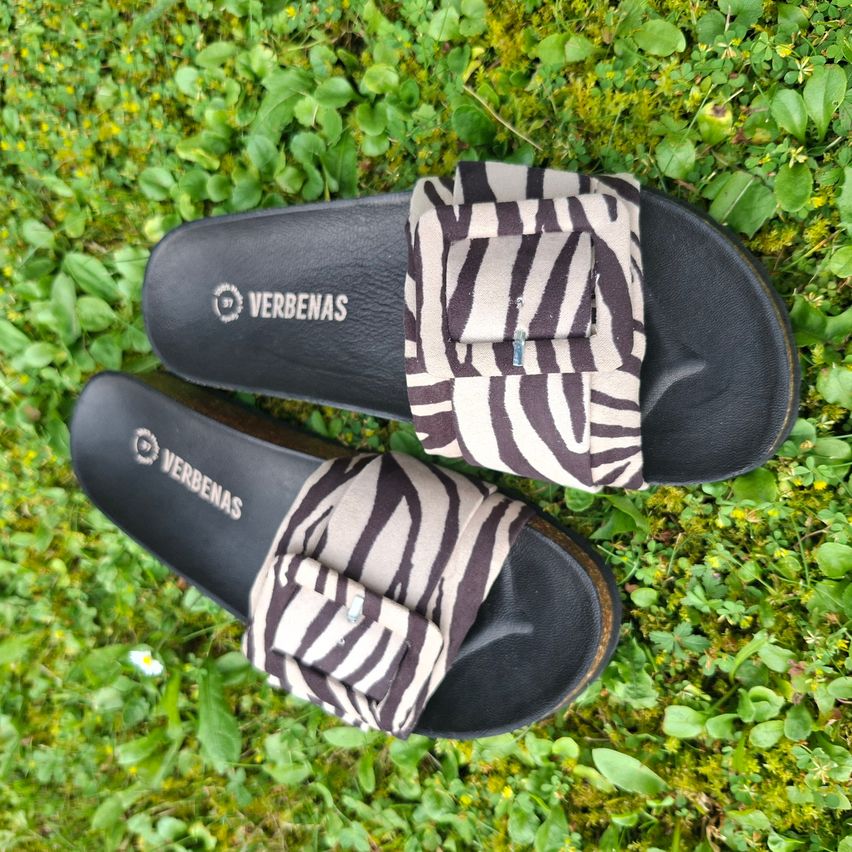 Ein Paar schwarze Verbenas-Schlappen mit Zebra-Print-Riemen liegen auf einer Grasfläche.