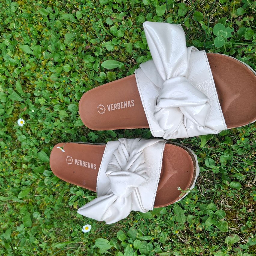 Ein Paar weiße Ledersandalen mit einer Schleifen-Dekoration liegen auf grünem Gras. Die Sohlen sind braun.