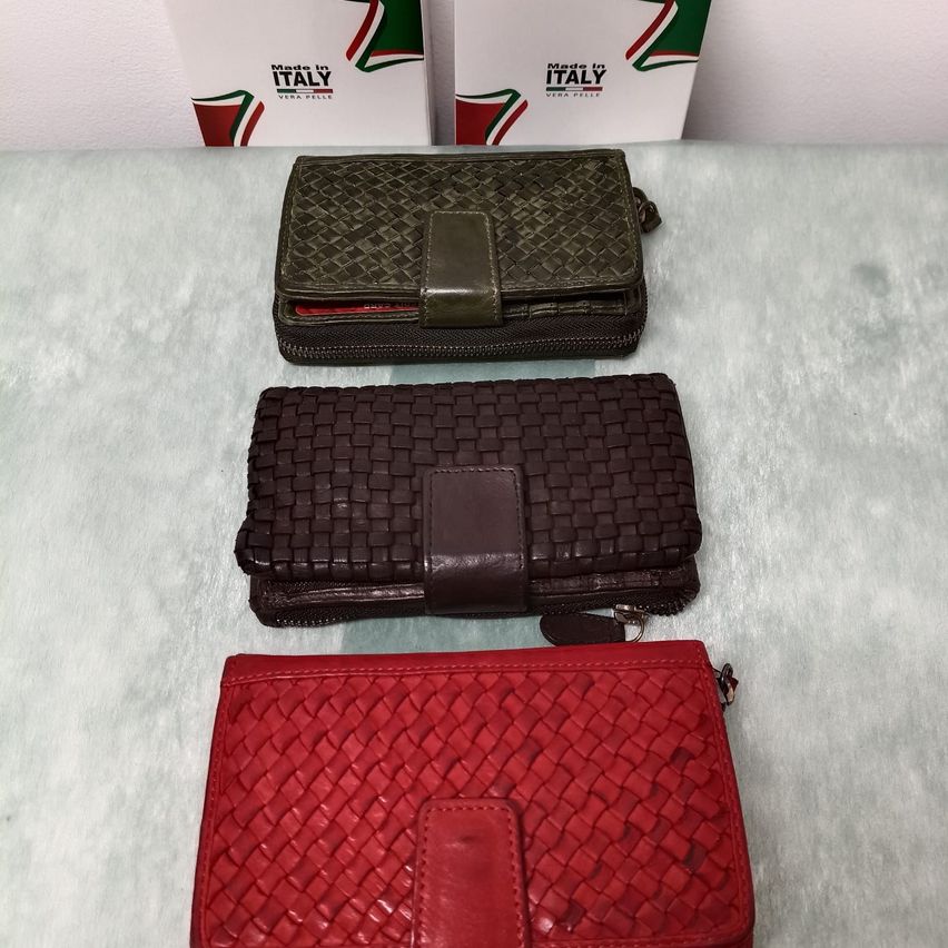 Drei Lederbrieftaschen in verschiedenen Farben liegen auf einer weißen Oberfläche. Die oberste Brieftasche ist grün, die mittlere braun und die unterste rot. Hinter den Brieftaschen befinden sich zwei weiße Schachteln mit grünen und roten Streifen.