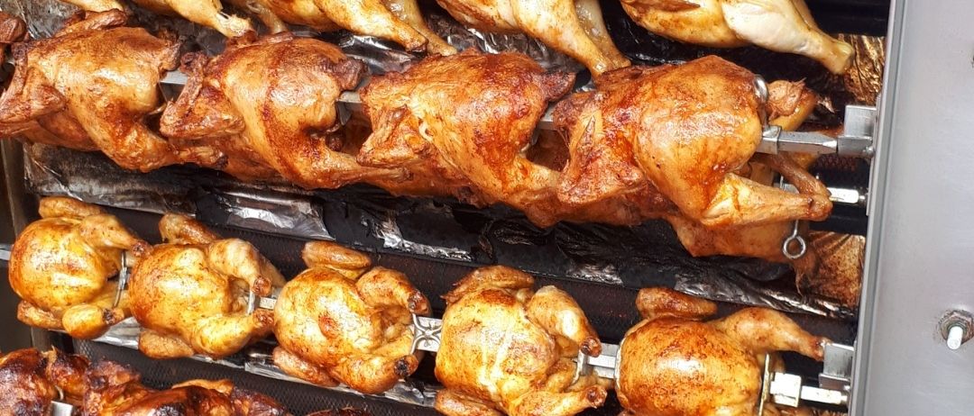 Mehrere Hühner werden in einem kommerziellen Drehspießofen gekocht, wobei ihre goldbraune Haut und knusprige Textur zu sehen sind. Der Ofen ist mit Regalen voller Hühner gefüllt, die jeweils an Metallhaken hängen.